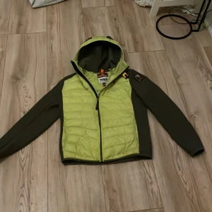 Grön och gul jacka från Parajumpers - Säljer en cool jacka från Parajumpers i grönt och gult. Jackan har en huva och dragkedja framtill. Den är designad med en patch på ärmen och har en quiltad look på framsidan. Perfekt för en sportig stil! Storlek y-m 