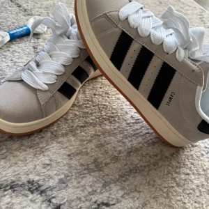 Adidas Campus sneakers i grått och svart - Snygga Adidas Campus sneakers i grå mocka med svarta ränder och vit sula. Skorna har klassisk snörning och en stilren design som passar perfekt till vardags. De är bekväma och lätta att matcha med olika outfits.