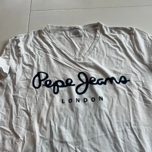 Vit t-shirt från Pepe Jeans - Säljer en vit t-shirt från Pepe Jeans med blå logotyp på framsidan. T-shirten har en v-ringad hals och är i slim passform. Perfekt för en avslappnad stil.