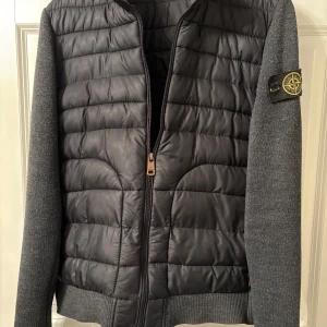 Grå jacka från Stone Island - Säljer en snygg grå jacka från Stone Island med quiltad front och stickade ärmar. Jackan har en dragkedja framtill och ett broderat märke på ärmen. Perfekt för höst och vår!
