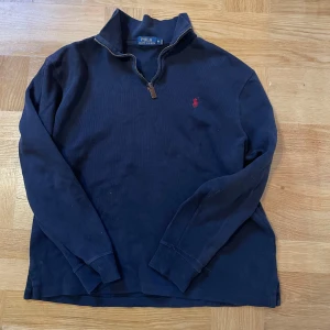 Mörkblå tröja från Polo Ralph Lauren - Säljer en stilren mörkblå tröja från Polo Ralph Lauren med dragkedja vid halsen och en liten röd logga på bröstet. Tröjan har långa ärmar och är perfekt för en avslappnad look.