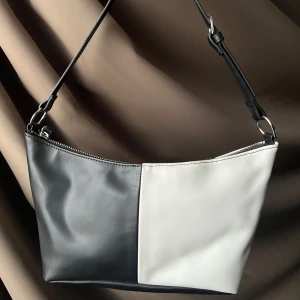Svart och vit axelväska - Snygg axelväska i svart och vitt med en stilren design. Väskan har en justerbar axelrem och en praktisk dragkedja upptill. Perfekt för att ge din outfit en modern touch.