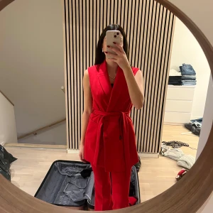 Röd väst med bälte - Snygg röd väst med knytbälte i midjan. Västen har en stilren design utan ärmar och en djup v-ringning. Perfekt för att ge en färgklick till din outfit.