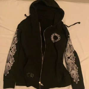 Svart hoodie från chrome hearts - Snygg svart hoodie med dragkedja och broderade vita mönster på ärmarna. Den har en huva med dragsko och ett tryck på bröstet. Perfekt för en cool och avslappnad stil.