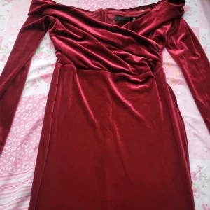 Vinröd sammetsklänning - Elegant vinröd långklänning i sammet med offshoulder-design. Klänningen har långa ärmar och en figurnära passform som ger en lyxig känsla. Perfekt för speciella tillfällen.