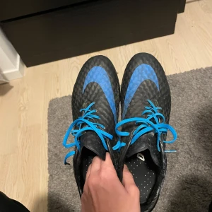 Svarta och blå fotbollsskor från Nike - Säljer ett par svarta och blå fotbollsskor från Nike med snörning. Skorna har en modern design med blå detaljer och Nike-logga på sidan. Perfekta för fotbollsplanen!