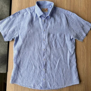 Blå kortärmad skjorta från Ströms - Bomull men likt linne i känslan. Säljer en stilren blå kortärmad skjorta från Ströms. Skjortan har en klassisk krage och knappar framtill. Perfekt för en avslappnad sommarlook. Den är tillverkad i ett lätt och bekvämt material. 