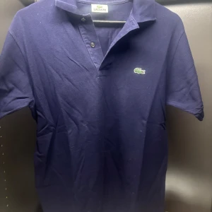 Mörkblå pikétröja från Lacoste - Säljer en klassisk mörkblå pikétröja från Lacoste med den ikoniska gröna krokodilen broderad på bröstet. Tröjan har korta ärmar och en stilren krage med knappar. Perfekt för en avslappnad och stilren look.