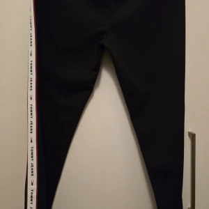 Svarta leggings från Tommy Jeans - Snygga svarta leggings från Tommy Jeans med röd och vit rand längs sidorna. Perfekta för en sportig look med en tight passform. Märkesloggan syns längs ränderna vilket ger en cool detalj.