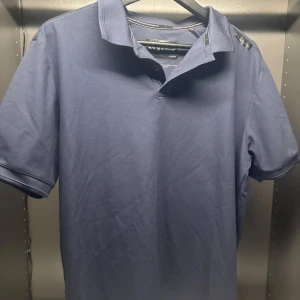 Blå pikétröja från Sail Racing - Snygg blå pikétröja från Sail Racing i storlek L. Perfekt för en avslappnad stil med klassisk krage och korta ärmar. Tillverkad med Sail Racing Performance Technology för extra komfort.