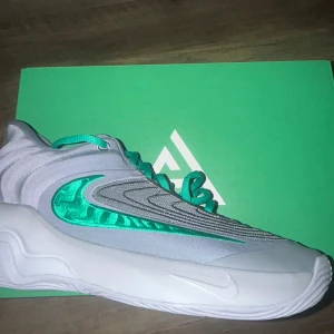 Nike Giannis sneakers i grått och grönt - Snygga Nike Giannis sneakers i grått med gröna detaljer. Skorna har en unik design med vågformade mönster och en tydlig Nike-logga. Perfekta för basket eller vardagsbruk. Kommer i originalkartong. Storlek 41 helt oanvända för små för mig som har legat och ett bra tag i paketet den kom i orginal pris 1100kr som sagt helt oanvända 