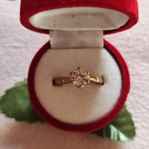 Guldfärgad ring med klar sten - Elegant guldfärgad ring med en stor klar sten i mitten. Ringen levereras i en vacker röd ask formad som en ros, perfekt för speciella tillfällen.