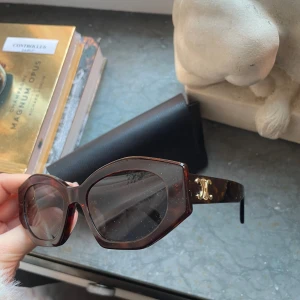 Bruna solglasögon från Celine - Snygga solglasögon från Celine i en klassisk brun nyans med en elegant fyrkantig form. Bågarna har en stilren design med gulddetaljer på sidorna som ger en lyxig touch. Perfekta för att höja din stil till nästa nivå. Fodral ingår