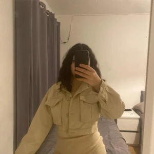 Beige kortklänning med jacka - Snygg beige kortklänning med en matchande jacka. Klänningen är tight och ärmlös, perfekt för en stilren look. Jackan har stora fickor och knappar framtill.