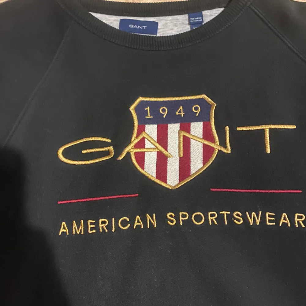 Säljer en svart sweatshirt från GANT med broderad logga och texten 'American Sportswear' på framsidan. Tröjan har långa ärmar och en rund halsringning. Perfekt för en sportig och stilren look.. Neuletakit & Villapaidat.