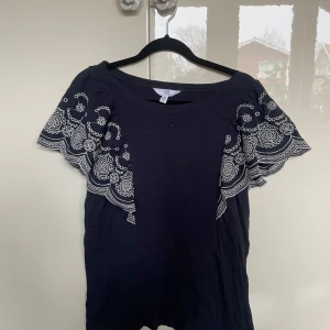 Mörkblå topp med spetsdetaljer - Snygg mörkblå topp spetsdetaljer på ärmarna. Toppen har en rund hals och korta ärmar med spetsmönster i vitt.