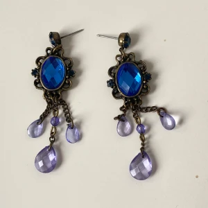 Blå örhängen med kedjor - Snygga örhängen med en stor blå sten i mitten och droppformade hängen. Designen inkluderar kedjor och en antik metallram som ger en vintagekänsla. Perfekta för att ge en färgklick till din outfit.