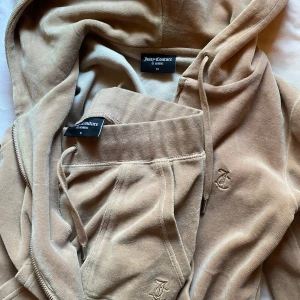 Beige velourset från Juicy Couture - Säljer ett snyggt beige velourset från Juicy Couture. Setet består av en hoodie med dragkedja och matchande byxor med snörning i midjan. Perfekt för en avslappnad stil med en touch av lyx. 🧥👖