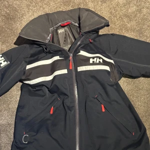 Mörkblå seglarjacka från Helly Hansen - Säljer en svart vindjacka från Helly Hansen med Helly Tech Protection. Jackan har dragkedja och vita detaljer samt en justerbar huva. 