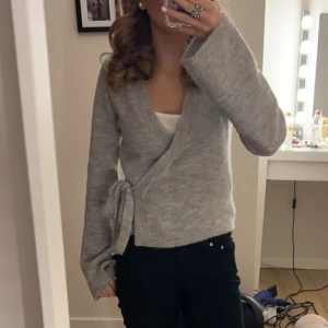Grå omlottkofta - Säljer en mysig grå omlottkofta med knytband i midjan. Koftan har långa ärmar och är perfekt för kyligare dagar. Den är både stilren och bekväm, vilket gör den till ett mångsidigt plagg i garderoben.