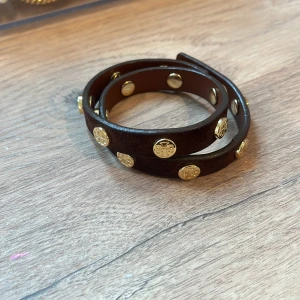 Brunt läderarmband med gulddetaljer - Snyggt brunt läderarmband med guldfärgade nitar. Armbandet är designat för att lindas runt handleden och har en stilren look. Perfekt för att ge en touch av elegans till din outfit.