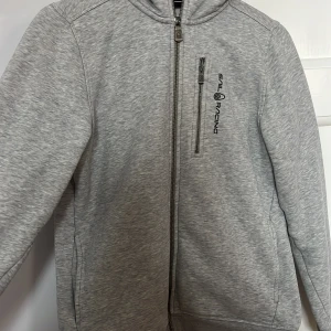 Grå hoodie från Sail Racing - Säljer en grå hoodie från Sail Racing med dragkedja och en liten bröstficka. Den har en logga på bröstet och en på huvan. Perfekt för en avslappnad stil.