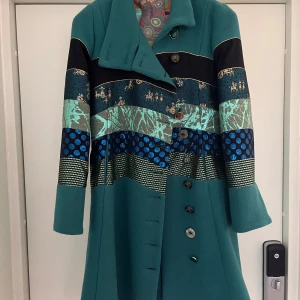 Turkos mönstrad kappa - Unik turkos kappa med en mix av mönster och färger. Den har långa ärmar och knappar framtill. Mönstren inkluderar prickar och abstrakta former i blått, svart och guld. Perfekt för den som vill sticka ut med sin stil.