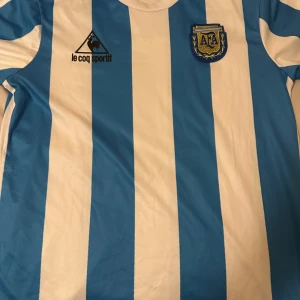 Argentina #10 maradona - Klassisk blå och vit randig fotbollströja från Le Coq Sportif med AFA-emblem på bröstet. Tröjan har korta ärmar och nummer 10 med texten 'Maradona' på ryggen. Perfekt för fotbollsfans och samlare!