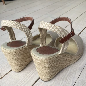 Beige kilklackssandaler med remmar - Beige kilklacksandaler med skimmer och flätad sula. Justerbara bruna remmar runt ankeln. Sköna! Storlek 38.