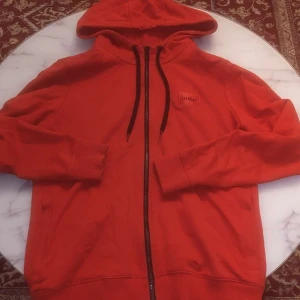 Röd hoodie från Hugo Boss - Säljer en snygg röd hoodie från Hugo Boss med dragkedja och justerbar huva. Perfekt för en casual look med stil. Den har en liten Hugo-logga på bröstet och ribbade muddar vid ärmslut och nederkant.