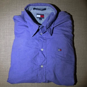 Lila skjorta från Tommy Hilfiger - Snygg lila skjorta från Tommy Hilfiger med klassisk krage och knappar framtill. Skjortan har en liten logotyp på bröstet och ett randigt mönster på insidan av kragen. Perfekt för en stilren look!