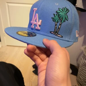 LA keps blå - Snygg blå keps från New Era med broderad LA-logga i rosa och palmmotiv. Perfekt för en cool och avslappnad stil. Kepsen har en platt skärm och är i storlek 7 (55,8 cm). 
