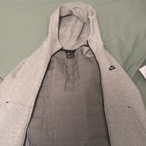 Grå quiltad jacka från Nike - Säljer en grå quiltad jacka från Nike med huva och dragkedja. Jackan har en stilren design med Nike-logga på bröstet. Perfekt för kyligare dagar.