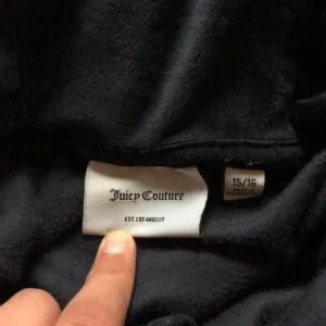 Svart velourjacka från Juicy Couture - Säljer en snygg svart velourjacka från Juicy Couture med huva och dragkedja. Jackan har glittriga detaljer med JC-logga på bröstet och Juicy Couture på ryggen. Perfekt för en stilren look!