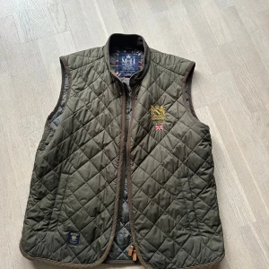 Olivgrön quiltad väst från Morris Gent's - Säljer en olivgrön quiltad väst från Morris Gent's. Västen har en dragkedja framtill och ett broderat emblem på bröstet. Perfekt för en stilren look.