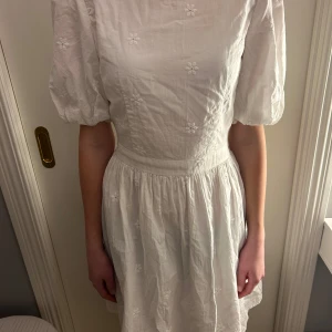 Vit klänning med blommönster - Charmig vit klänning med blommönster och puffiga kortärmar. Klänningen har en öppen rygg med knytning och en luftig kjol. Perfekt för sommarens varma dagar.