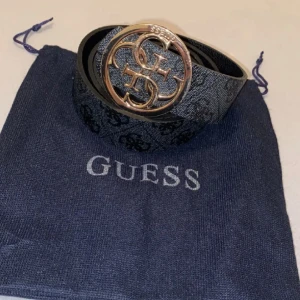 Svart bälte från Guess - Säljer ett snyggt svart bälte från Guess med en stor, rund guldspänne som har Guess-loggan. Bältet är i skinn och kommer med en praktisk förvaringspåse. Perfekt för att ge en stilren touch till din outfit! ✨