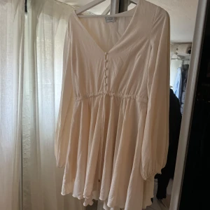 Vit klänning från nakd  - Säljer en söt vit klänning i storlek 34. Klänningen har en v-ringad design med knappar framtill och långa ärmar. 