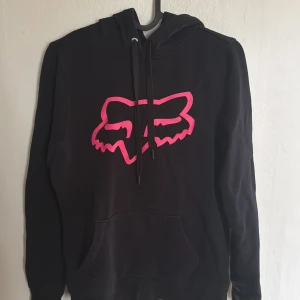 Svart hoodie från Fox Racing - Säljer en svart hoodie från Fox Racing i bra skick. Den har en stor rosa logga på framsidan och en praktisk ficka. Perfekt för höst och vinter med sin mysiga känsla. Huvan har justerbara snören för extra komfort. Passar både till vardags och chillkvällar. Priset kan diskuteras.