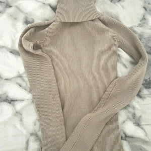 Beige ribbad polotröja - Säljer en supermysig beige polotröja i ribbat material. Perfekt för kyliga dagar med sin långa ärmar och höga krage. Tröjan är både stilren och bekväm, idealisk för höst och vinter. Passar perfekt till jeans eller kjol för en avslappnad look.