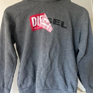 Grå hoodie från Diesel - Säljer en grå hoodie från Diesel i mycket bra skick. Den har en stor logga i rött och svart på framsidan. Perfekt för höst och vinter, superbekväm och stilren. Passar både till vardags och chillkvällar. 🖤