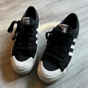 Svarta sneakers från Adidas Nizza - Säljer ett par snygga svarta Adidas sneakers i mycket bra skick. De har den klassiska tre-randiga designen i vitt på sidan och en vit tåhätta. Perfekta för vardagsbruk eller en avslappnad stil. Skorna har snörning och är bekväma att gå i. Passar perfekt till jeans eller shorts!