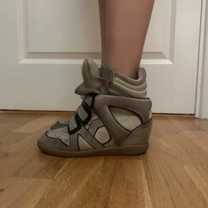 Beige sneakers med kilklack - Säljer ett par super fina beiga isabel marant skor med svarta detaljer, relativt bra skick men har används en del. Några slitningar på sulan under och i skon (dom syns eller känns inte dock) några synliga slitningar vid kardborren men annars riktigt snygga. Jätte coola skor som är efterfrågade hos många och trender mycket. Tyvärr är dom nu för små för mig💞💞dom är i storlek 37, skriv för fler bilder om så önskas!☺️pris kan även diskuteras🥰🥰