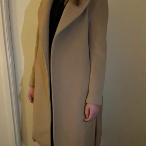 Beige kappa med bälte - Säljer en elegant beige kappa i mycket bra skick. Den har en stilren design med ett knytbälte i midjan och långa ärmar. Perfekt för höst och vår när du vill hålla dig varm och snygg. Passar både till vardags och festliga tillfällen. Helt oanvänd från hm!! Perfekt till hösten.
