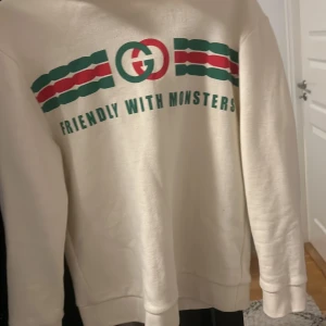 Vit hoodie från Gucci - Säljer en snygg vit hoodie från Gucci med texten 'Friendly with Monsters' och coola ränder i grönt och rött. Den är i bomull och har en klassisk passform med långa ärmar. Perfekt för både vardag och chill.