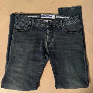 Gråa jeans från Jacob Cohen - Säljer ett par snygga svarta jeans från Jacob Cohen i storlek 34. De är i mycket bra skick och har en klassisk femficksdesign med knappgylf. Dock lite sliten batch där bak. Perfekta för både vardag och fest!