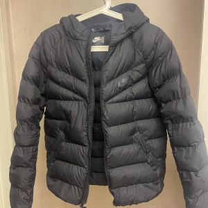 Svart dunjacka från Nike - Säljer en svart dunjacka från Nike i storlek 158. Jackan har en dragkedja, fickor och varm huva. Den har använts ganska flitigt men är i gott skick. Jackan ser nästan grå ut på korten, men den är svart i verkligheten. Köpt på Stadium och passar både tjej och kille.