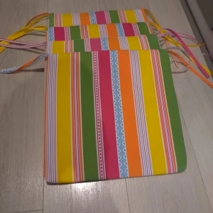 Färgglad randig sittdyna 35×35 cm - Säljer en superfin och färgglad sittdyna med ränder i grönt, rosa, gult, orange och blått. Perfekt för att liva upp uteplatsen eller balkongen. Dynan har knytband för enkel fastsättning. Den är i mycket bra skick och redo för sommarens alla sittningar!