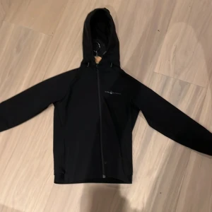Svart jacka med huva - Säljer en svart jacka från sail racing i  bra skick. Den har en praktisk huva och dragkedja framtill. Perfekt för höst och vår, med en stilren design som passar till det mesta. Jackan har långa ärmar och en liten logga på ärmen. Perfekt för dig som vill ha en snygg och funktionell jacka!Det är storlek 150.org pris 1499kr