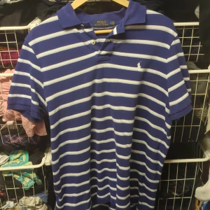 Blå och vit randig pikétröja från Ralph Lauren - Säljer en klassisk blå och vit randig pikétröja från Ralph Lauren. Den har en broderad logga på bröstet och är i mycket bra skick. Perfekt för både vardag och festliga tillfällen. Passar bra till jeans eller chinos för en stilren look.